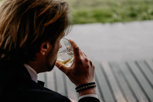 a courageous brutal man dressed in a suit with a glass of strong alcohol and a cigarette in his hands sits in the backyard of his home - 写真・画像