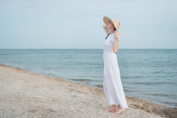 Young woman in long white dress and hat standing on beach. Evening sea - Foto, imagen