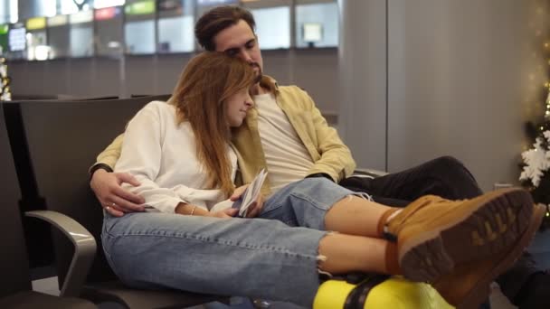 Tired young caucasian couple sitting and trying to sleep in departure lounge with suitcase and waiting for delayed flight. Stressed and exhausted tourists waiting for their flight embraced - Imágenes, Vídeo