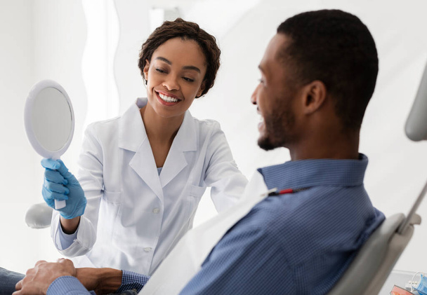 Mooie zwarte tandarts houden spiegel voor mannelijke patiënt - Foto, afbeelding