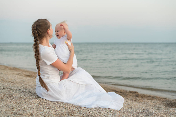 Young mother sitting on beach and holding baby. Joys of motherhood. - Фото, зображення