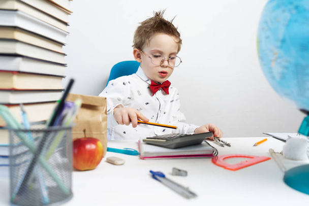 Back to school: a little boy in a shirt and glasses sits at his desk and does the calculations on a calculator. Portrait. - Φωτογραφία, εικόνα