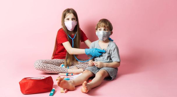 Personal protective equipment: a girl and a masked boy are playing q doctor. The girl uses a stethoscope and listens to the boy s breath. Portrait on a pink background. - Foto, Imagem