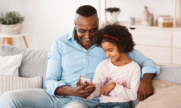 Reifer afroamerikanischer Mann und seine Enkelin nutzen Smartphone, spielen Videospiele oder surfen im Internet - Foto, Bild