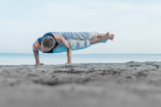 Young flexible man standing on hands in yoga position on the seashore beach - Fotoğraf, Görsel