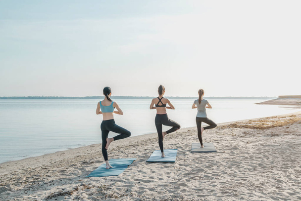 Rear view shot of women group practicing healthy lifestyle, yoga on beach. - Photo, image