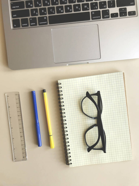 Az asztalon: egy notebook egy ketrecben, fekete szemüveg, színes tollak és egy vonalzó. a közelben van egy laptop - Fotó, kép