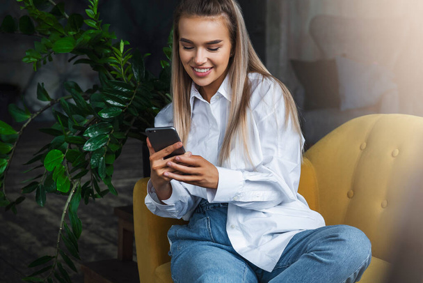 Smiling beautiful blonde girl sits on yellow sofa and holds smartphone. Millennial woman communicates with friends - Photo, image