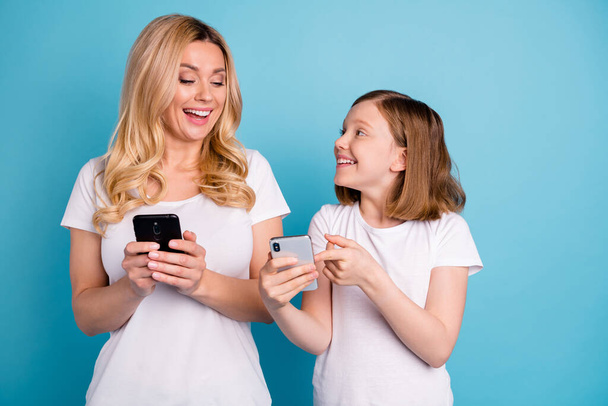 Photo of two people beautiful mom lady little daughter excited mood spend leisure hold telephones showing funny post blog screen wear casual white s-shirts isolated blue color background - Foto, Bild