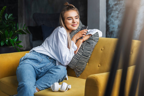 Girl enjoying being home, sitting on sofa, holding smartphone in hand. Young female using mobile device for fun - Fotoğraf, Görsel