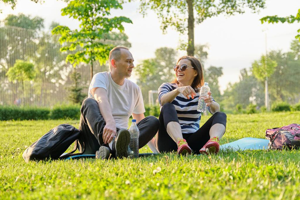 Middle-aged couple sitting on yoga mat, man and woman talking relaxing drinking water. Active healthy lifestyle, relationship, sport, fitness in mature people - Фото, изображение