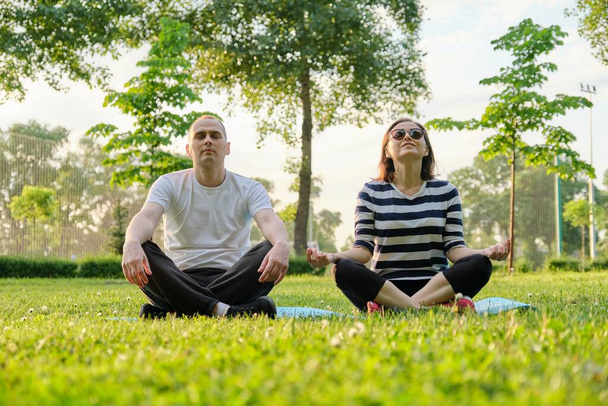 Çift, erkek ve kadın, parkta nilüfer pozisyonunda oturmuş meditasyon yapıyorlar. Yoga, fitness, orta yaşlı insanlarda aktif sağlıklı yaşam tarzı - Fotoğraf, Görsel