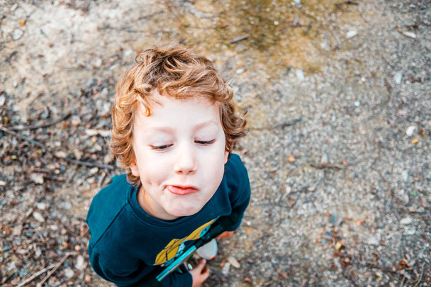 Blond boy making funny gestures and faces photographed from above with natural ground background. - Photo, Image