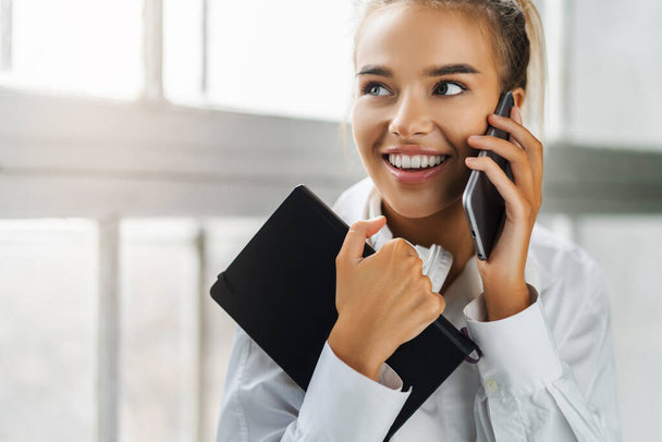 Smiling girl talking on smartphone, holding black notepad. Beautiful girl use mobile phone for work, call clients - Foto, afbeelding