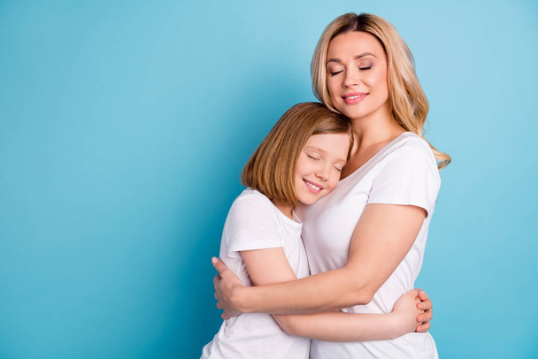Φωτογραφία προφίλ δύο ανθρώπων νεαρή όμορφη μαμά κυρία μικρή κόρη αγκαλιάζει τα μάτια κλειστά κρατώντας τα χέρια στοργική φορούν casual λευκό s-shirts απομονωμένο φόντο μπλε χρώμα - Φωτογραφία, εικόνα