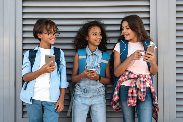 Crianças da escola sorrindo usando smarthphones e assistindo no telefone celular na rua em pé sobre fundo cinza
 - Foto, Imagem