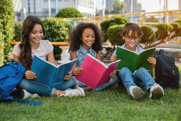 Lindos niños felices de la escuela con mochilas y cuadernos sentados en el parque de hierba al aire libre
 - Foto, Imagen