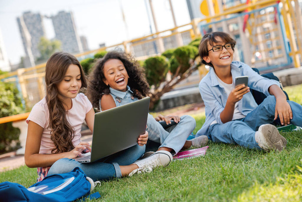 Group of multiethnic elementary school children sitting on playground grass looking at laptop computer - Foto, afbeelding