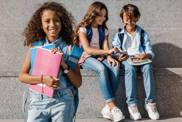 Portrait photo de mignonne petite écolière souriant et regardant la caméra avec des enfants de l'école, sacs à dos et cahiers sur fond extérieur
 - Photo, image