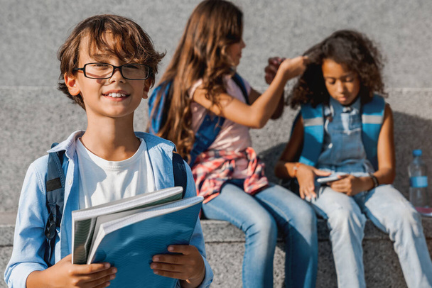 Портрет маленького школяра в окулярах посміхається і дивиться на камеру з однокласниками на фоні на відкритому повітрі
 - Фото, зображення