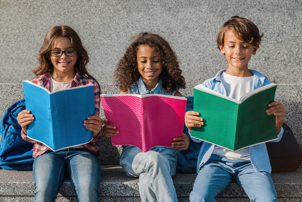 Primer plano de niños sonrientes de la escuela con mochilas sosteniendo cuadernos y divirtiéndose al aire libre
 - Foto, imagen