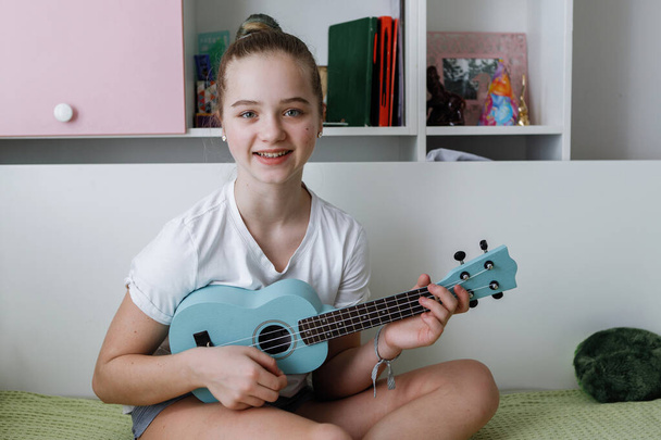 寝室のベッドに座っている間、青いウクレレギターで髪のパンを持つ十代の女の子が演奏します。家具. - 写真・画像