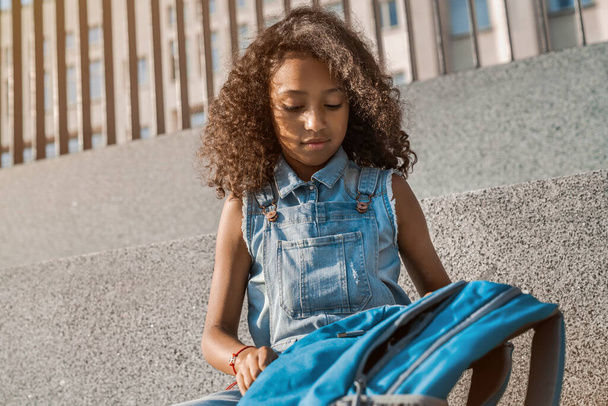 Portrait of cute little girl with school backpack sitting outdoors at school yard - 写真・画像