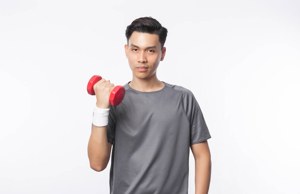 Young Asian sport man in grey t-shirt and white wristband doing exercise with dumbbell and looking camera isolated over white background. - Фото, изображение