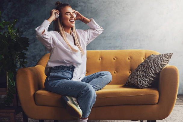 Beautiful blonde woman in white headphones sits on yellow sofa, enjoys listening to podcast. Relaxing girl in shirt - Foto, Imagem