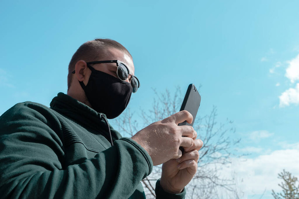 Young man wearing face mask. Handsome man in sunglasses wear black medical mask holding smartphone, sky background, copy space. Pandemic coronavirus covid-19 quarantine period concept. - Photo, Image
