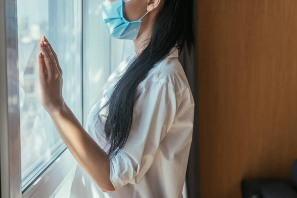 cropped view of young woman in medical mask standing near window at home - Fotoğraf, Görsel