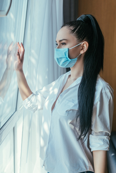 sad girl in medical mask looking through window at home - Photo, Image