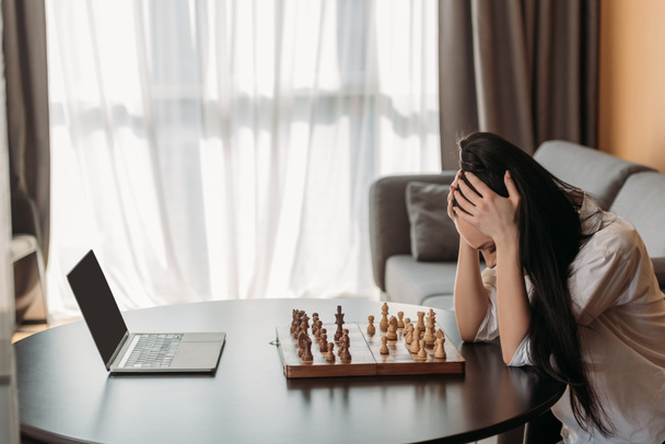 depressed woman touching bowed head while sitting near laptop with blank screen and chessboard - Photo, Image