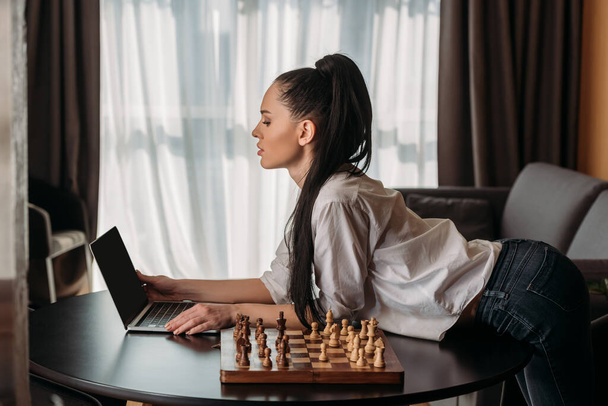 side view of attractive woman looking at laptop with blank screen near chessboard on table - Fotoğraf, Görsel