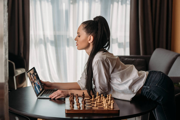 Seitenansicht der schönen Frau mit Videoanruf mit Freund in der Nähe Schachbrett auf dem Tisch - Foto, Bild