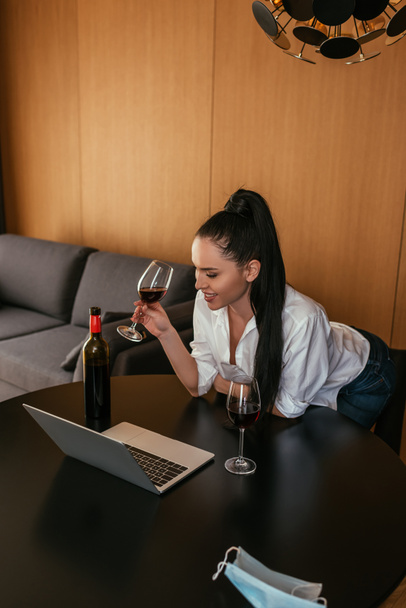 uśmiechnięta młoda kobieta trzymająca kieliszek czerwonego wina podczas wideo czatu na laptopie - Zdjęcie, obraz
