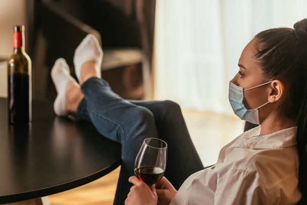 selective focus of depressed young woman in medical mask holding glass of red wine while sitting with legs on table - Fotó, kép