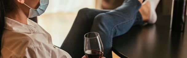 Ausgeschnittene Ansicht einer Frau in medizinischer Maske, die ein Glas Rotwein hält, während sie mit Beinen auf dem Tisch sitzt, horizontales Bild - Foto, Bild