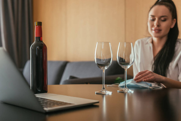 foco seletivo da mulher sentada à mesa perto de copos de vinho, garrafa, máscara médica e laptop
 - Foto, Imagem