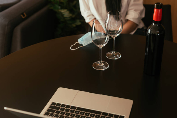 vista cortada da mulher sentada à mesa perto de copos de vinho, garrafa, máscara médica e laptop
 - Foto, Imagem