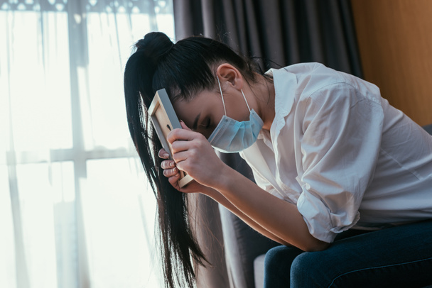 καταθλιπτική νεαρή γυναίκα με ιατρική μάσκα θρηνεί ενώ κάθεται με σκυμμένο κεφάλι και κρατώντας κορνίζα - Φωτογραφία, εικόνα