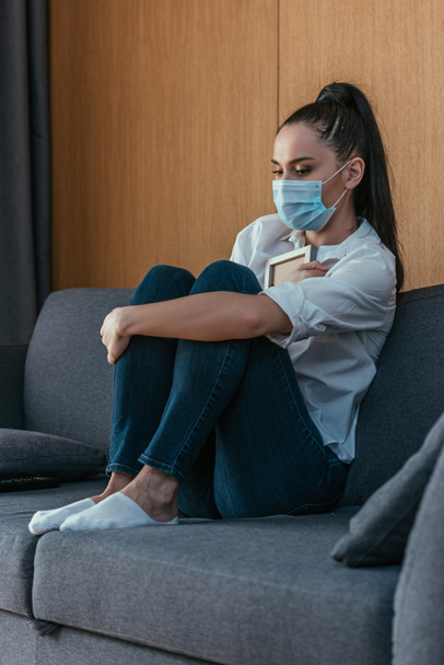mujer joven deprimida en máscara médica sosteniendo marco de fotos cerca del pecho mientras está sentado en el sofá
 - Foto, Imagen