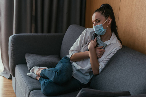 stressé jeune femme dans le masque médical tenant cadre photo près de la poitrine tout en étant assis sur le canapé
 - Photo, image