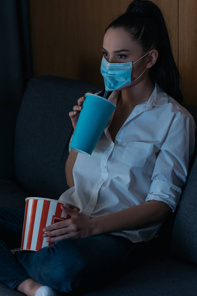 young woman in medical mask holding soda and popcorn while watching tv at home - Foto, Imagem