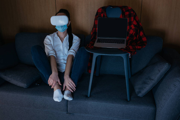 depressieve vrouw zitten op de bank en met behulp van vr headset in de buurt van laptop met een leeg scherm op stoel - Foto, afbeelding