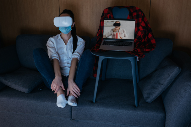 young woman sitting on sofa near laptop with boyfriend eating popcorn while using vr headsets together - Foto, Imagen