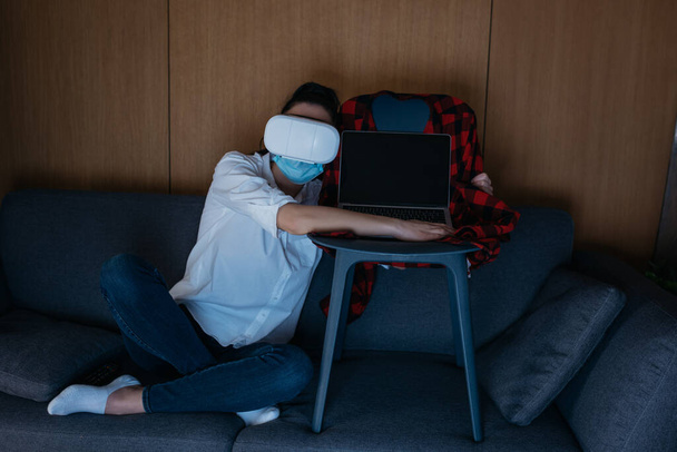 mujer joven en auriculares vr sentado en el sofá y abrazando el ordenador portátil con pantalla en blanco en la silla
 - Foto, Imagen