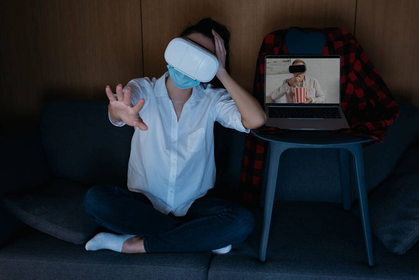 νεαρή γυναίκα outstretching χέρι, και ο φίλος στην οθόνη του φορητού υπολογιστή τρώει ποπ κορν, ενώ χρησιμοποιώντας ακουστικά VR μαζί - Φωτογραφία, εικόνα