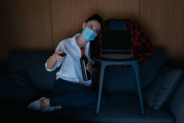 young woman in medical mask holding bottle of wine and tv remote controller while sitting on sofa near laptop with blank screen - Fotoğraf, Görsel