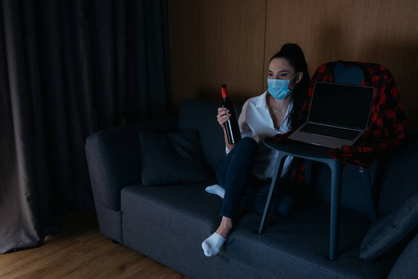 jeune femme dans le masque médical tenant bouteille de vin tout en étant assis sur le canapé près d'un ordinateur portable avec écran blanc
 - Photo, image